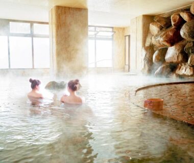 Steaming indoor onsen bath with rock wall, large windows, two guests relaxing in shallow thermal water at Hotel Shinko, Yamanashi