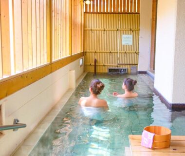Two guests relaxing in indoor onsen with wooden bucket and windows at Ooedo Onsen Monogatari Hotel Shinko, Yamanashi