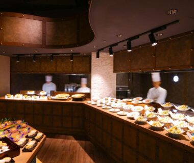 Chefs arranging buffet plates along curved wooden counter in dining area at Ooedo Onsen Monogatari Premium Atami, Shizuoka