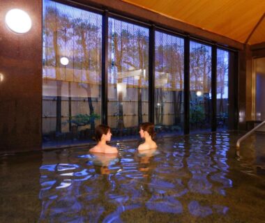 Guests soaking in a spacious indoor onsen with wooden ceiling and large windows at Ooedo Onsen Monogatari Premium Atami, Shizuoka
