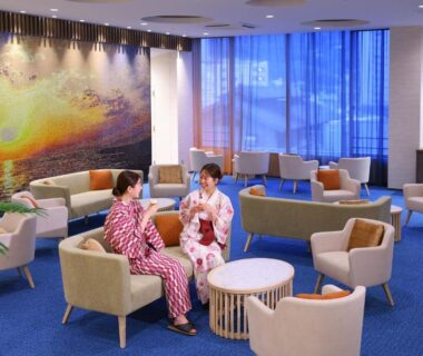 Guests in yukata conversing in a spacious ryokan lounge with mural, armchairs, low tables and large windows
