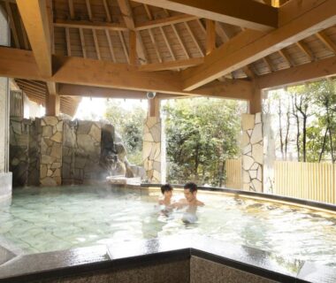 Guests relaxing in a spacious roofed onsen with wooden beams, stone walls and garden view at Kaga ryokan in Ishikawa