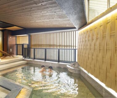 Two guests soaking in indoor onsen with wooden ceiling and bamboo-textured wall at Ooedo Onsen Monogatari Kaga, Ishikawa