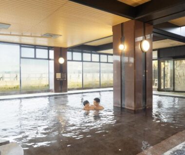Indoor communal onsen bath with large windows and two bathers at Ooedo Onsen Monogatari Premium Kaga Maruya in Ishikawa