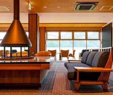 Spacious lounge with central fireplace, wooden seating and large windows overlooking greenery at Ooedo Onsen Monogatari Kinosaki in Hyōgo