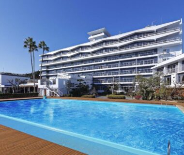 Large outdoor pool and deck before a white multi-story ryokan building with palm trees in Wakayama