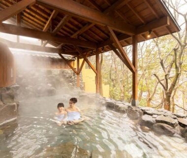 Steaming covered outdoor onsen with wooden roof, stone edging and tree-filled backdrop at Ooedo Onsen Monogatari Yamanaka