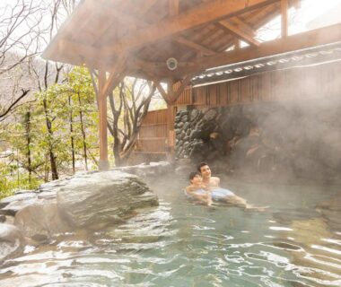 Guests relaxing in a steaming outdoor onsen with wooden shelter and surrounding trees at a ryokan in Ishikawa