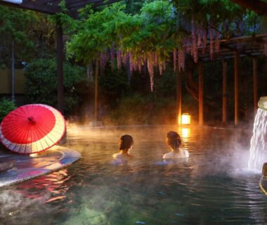 Steaming outdoor onsen with two bathers, red paper umbrella, lanterns and stone waterfall at Ooedo Onsen Monogatari Shinshu Kakeyu Nagano