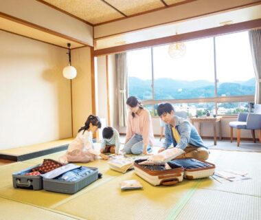 Family relaxing on tatami floor with open suitcase and mountain view at Ooedo Onsen Monogatari Ureshinokan, Saga