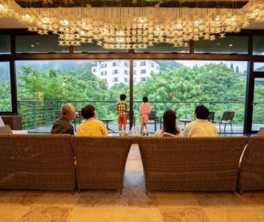 Guests seated in lounge facing large window overlooking trees and hotel building at Ooedo Onsen Monogatari Yamanaka