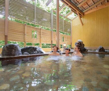 Guests relaxing in indoor onsen with bamboo blinds and stone edging at Ooedo Onsen Monogatari Yamanaka Saichoraku, Ishikawa