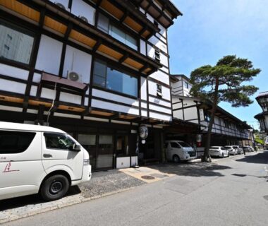 Row of traditional multi-story ryokan buildings with wooden beams, parked vans and pine tree along a sunlit street in Gunma