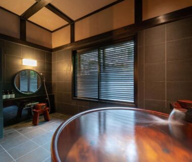 Round wooden soaking tub in tiled bath with vanity mirror and window blinds at Osawayama Onsen Osawakan, Niigata