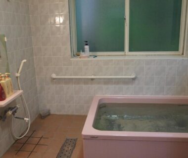 Tiled bathroom with pink bathtub, handheld shower, wall grab bar, frosted window and toiletries on a small shelf