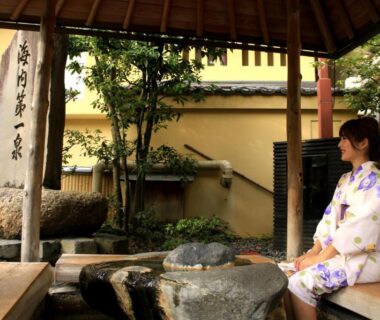 Guest in yukata seated at covered stone footbath with wooden posts and garden at Ougiya Ryokan in Hyōgo