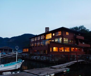 Lit wooden ryokan building on waterfront with docked fishing boats at dusk in Mie, Kansai