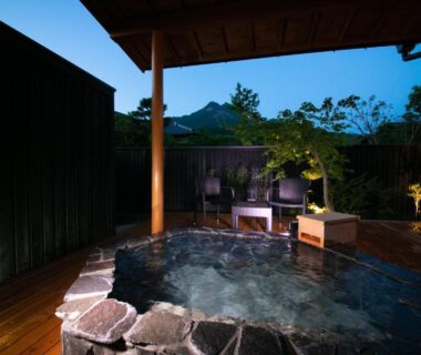 Stone outdoor onsen on wooden deck with mountain view and evening lantern lighting at Oyado Den Rikyu, Ōita