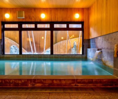 Indoor communal hot spring bath with wooden walls, stone seating and large windows at Oyado Koto No Yume, Gifu
