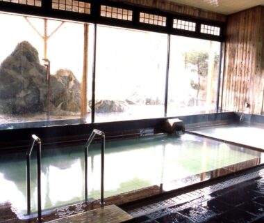 Large indoor onsen with tiled bath, handrails and rock garden visible through windows at Oyado Sansui Nagano