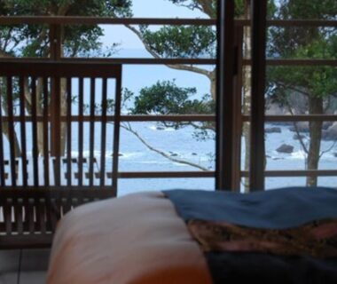 Low bed and wooden chair opening to balcony with sea, rocky shoreline and pine trees visible beyond