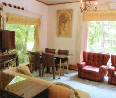 Bright sitting area with sofas, dining table and large garden-view windows at Pension Hakuba 70 Nagano