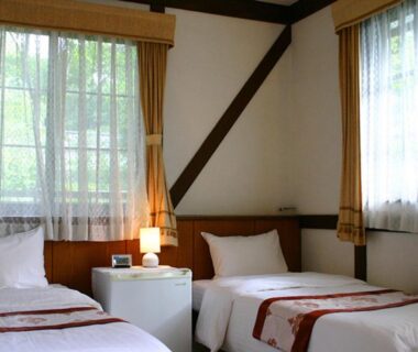 Light-filled twin bedroom with two beds, bedside table, lamp and curtained windows at Pension Marie in Yamanashi