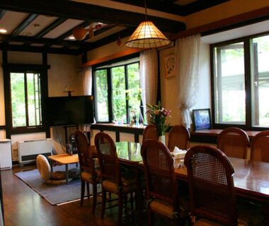 Wooden dining table and chairs in Pension Marie common room, windows showing green foliage outside