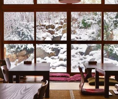 Low tables and zabuton on tatami facing a snow-covered garden through wooden-framed windows at Plum House Otaru