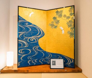 Gold folding screen with blue wave motif on wooden shelf beside lamp in ryokan interior