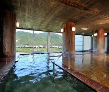 Indoor onsen bath with wooden pillars and large windows overlooking river and mountains in Nagano