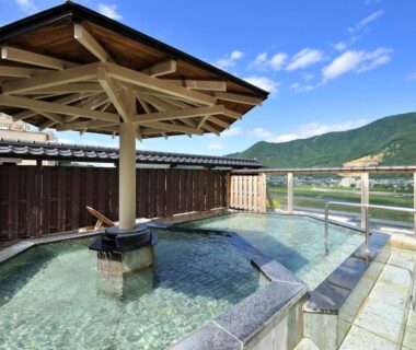 Outdoor onsen bath with stone edging, wooden pergola and glass railing overlooking river and distant mountains in Nagano
