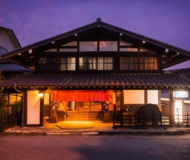 Warmly lit traditional wooden entrance with red noren and shoji windows at twilight, Ryokan Asunaro, Gifu