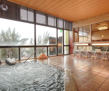 Tiled indoor onsen with wooden ceiling, washing stools and floor-to-ceiling windows overlooking forested hills