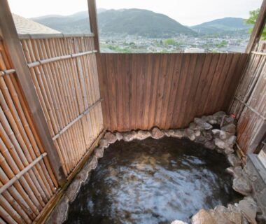 Stone-lined outdoor onsen with bamboo screening and distant mountain town view at Ryokan Fukinoya in Oita