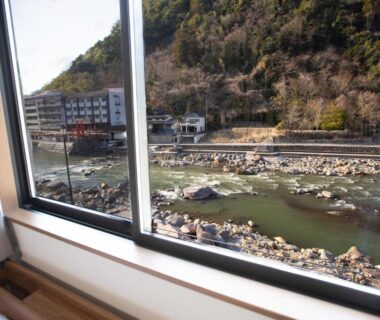 River view through ryokan room window with windowsill bench, rocky riverbank and hillside buildings in Ōita