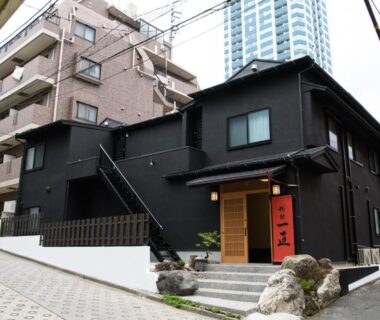 Two-story black ryokan exterior with red entrance curtain and stone steps in urban Tokyo neighborhood