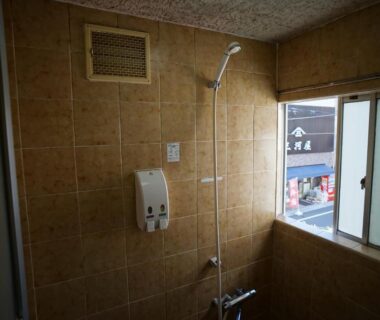 Compact tiled shower stall with wall soap dispenser and window overlooking street at Ryokan Katsutaro Tokyo
