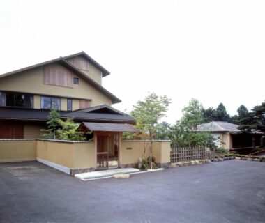 Multi-story traditional ryokan exterior with entrance gate, low wall and planted shrubs in Kanagawa