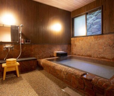 Stone-lined indoor onsen bath with wooden walls, shower station and stool at Ryokan Misato, Kumamoto, Kyūshū
