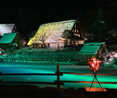Thatched houses lit at night beside a snow-lined river, lantern in foreground, Ryokan Murayama, Chūbu, Gifu, Japan