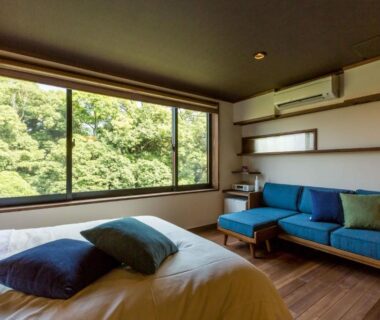 Guest room with bed, blue sofa, wooden floor and large window overlooking green trees in Saga