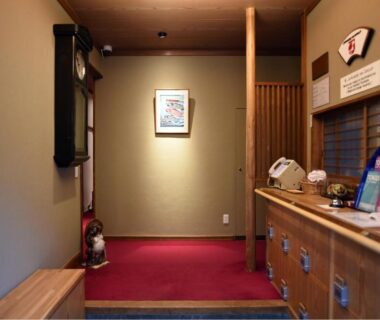 Small ryokan reception with wooden counter, brochure display, red carpet entry and framed artwork at Ryokan Sansuiso in Tokyo