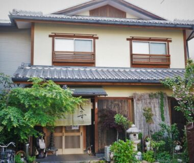 Two-story ryokan facade with noren, tiled roof, wooden entrance and potted greenery in Kyoto neighborhood