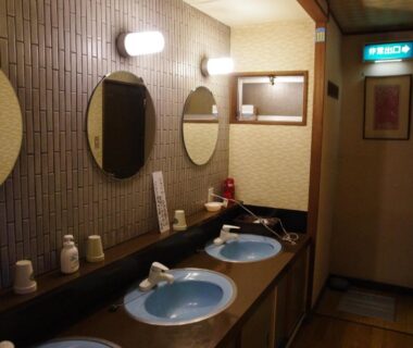 Narrow ryokan wash area with twin blue basins, round mirrors, wooden counter and warm wall lighting at Ryokan Takayama, Gifu