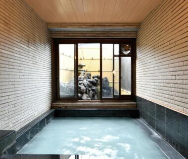 Indoor onsen bath with wooden ceiling, stone-lined tub, tiled walls and a window at Ryokan Tamura, Gunma