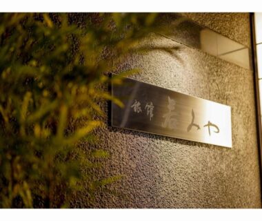 Metal nameplate with Japanese characters on textured wall illuminated by warm light, bamboo leaves in foreground