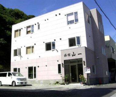 Three-story ryokan exterior with entrance sign, parked car and surrounding hillside in Hokkaidō, Japan