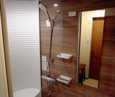 Compact wooden-paneled bathroom with handheld shower, mirror and shelves at Ryokan Yamamuro in Ishikawa, Chūbu, Japan