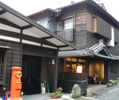 Exterior of traditional wooden ryokan with tiled roof, lantern-lit entrance and red post box on narrow street in Oita